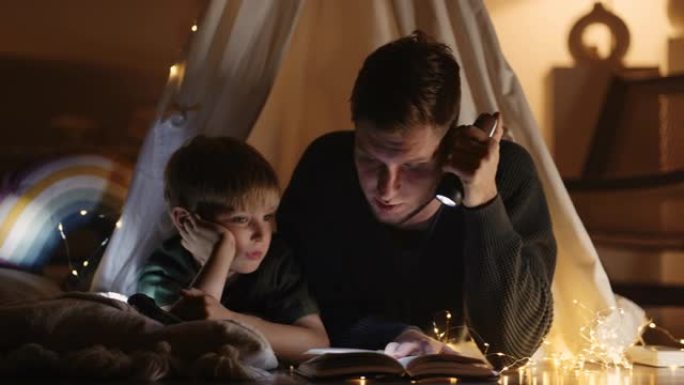 爸爸和小儿子拿着手电筒，看书，晚上躺在黑暗房间里用灯光装饰的帐篷里聊天和笑