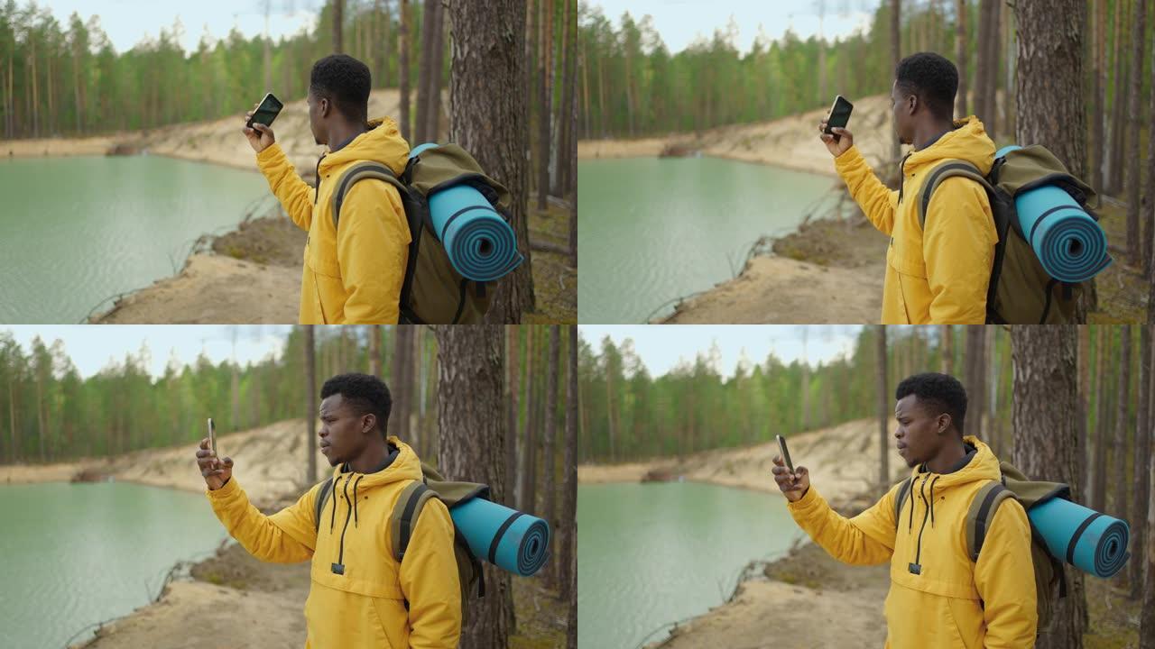 一个背着背包的黑人在湖对面的山林中徒步旅行时使用手机。拍照和博客