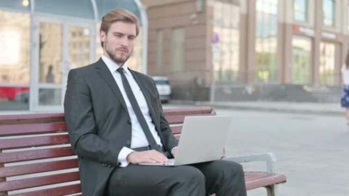 商人坐在户外的长凳上，用笔记本电脑看着相机