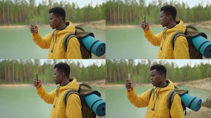 一个黑人正走在山顶上，在智能手机上输入消息和照片。他肩上有一个大背包。在山里徒步旅行。山顶