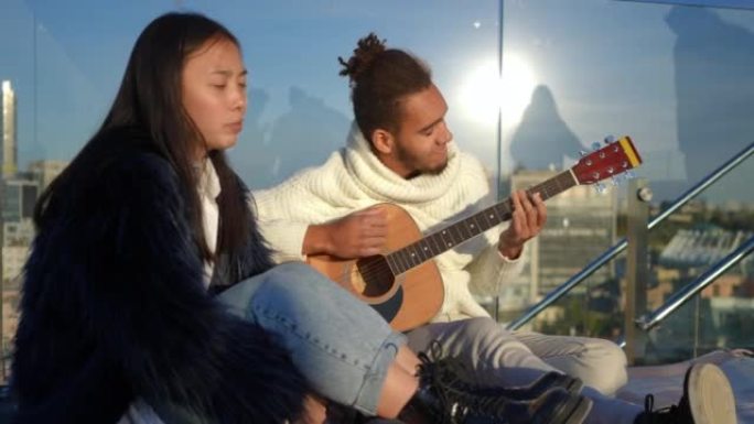 微笑的男人弹吉他的肖像，女人在日落时坐在城市的屋顶上唱歌。热爱无忧无虑的非洲裔美国人和亚洲夫妇周末喜