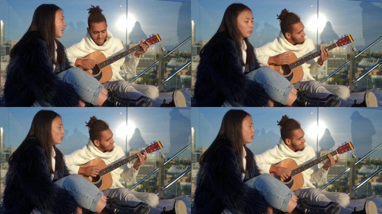 微笑的男人弹吉他的肖像，女人在日落时坐在城市的屋顶上唱歌。热爱无忧无虑的非洲裔美国人和亚洲夫妇周末喜