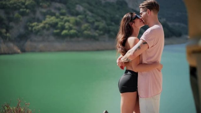 可爱的年轻夫妇站在美丽的绿松石湖边拥抱在一起