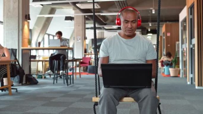DS男子坐在办公办公室的秋千上，戴着红色耳机在笔记本电脑上播放视频