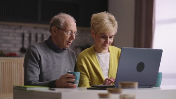 两名退休人员正在家里的厨房里使用笔记本电脑，上网冲浪，网上购物，老人和女人正在观看展出的图片