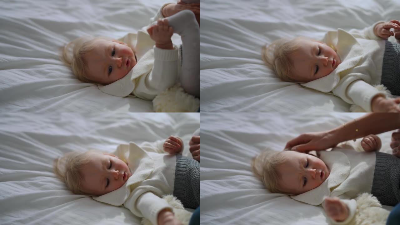 可爱的放松婴儿躺在床上的肖像，无法辨认的女人用小脚抚摸脸颊和鼻子，并以慢动作抚摸着脸。无忧无虑的高加