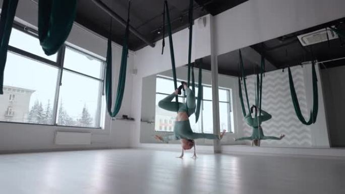 年轻微笑的女人练习空气拉伸秋千。健身俱乐部绿色吊床空中飞行瑜伽练习