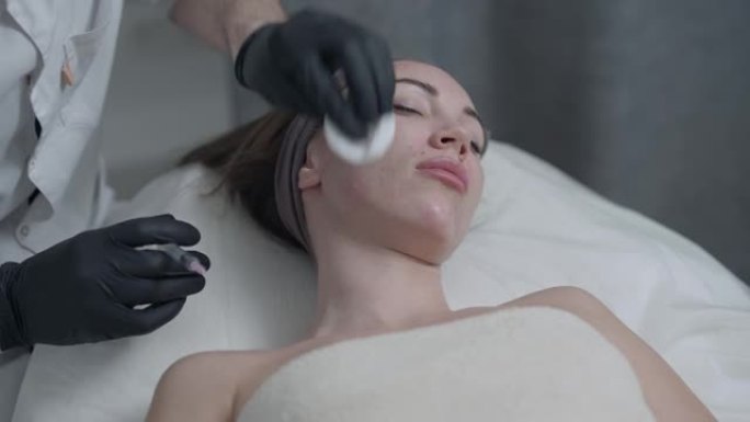 年轻苗条的白人妇女的肖像躺在美容院，作为专业美容师，用慢动作注射器在颈部皮肤注射肉毒杆菌填充剂。抗衰
