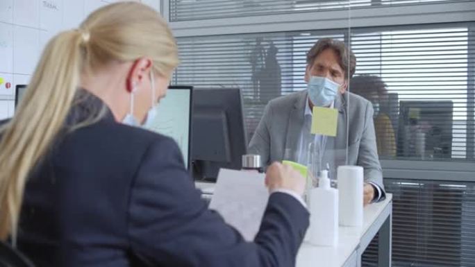 两名员工在办公室戴着口罩在办公桌前的防护屏上交谈