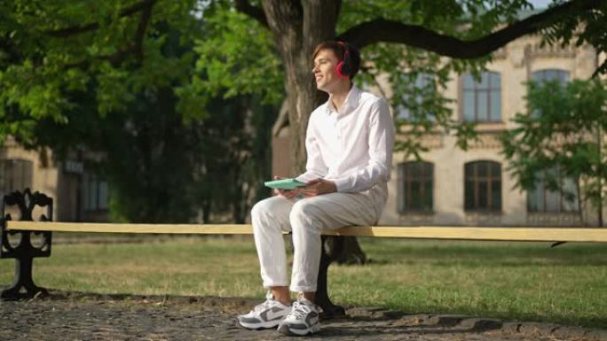 宽镜头快乐轻松的高加索年轻人坐在阳光明媚的大学院子里的长凳上，戴着耳机听音乐唱歌和微笑。快乐学生在户