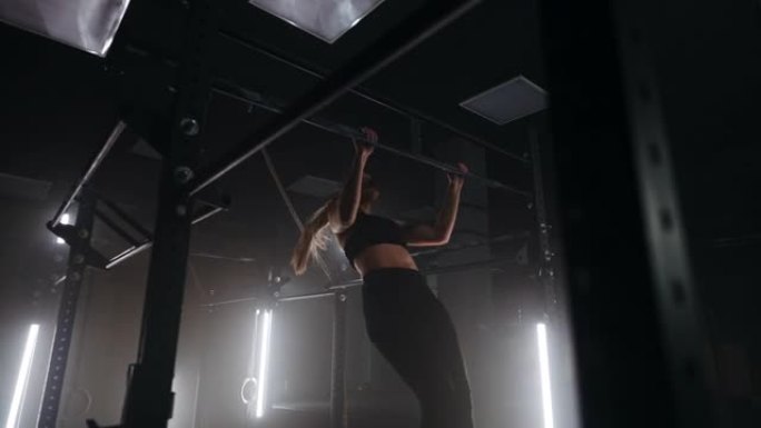 在昏暗的体育馆里，一名年轻女子在漂亮的霓虹灯背光照射下，把身子靠在单杠上。耐力和毅力拉起运动的目标