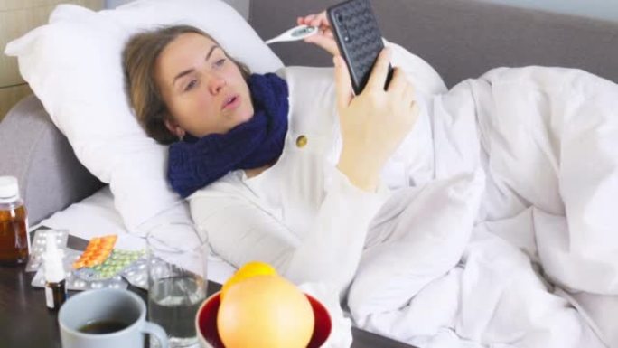 患有重感冒，流感或冠状病毒的患病年轻女子发热拿着温度计，同时通过智能手机通过视频链接咨询医生。生病的