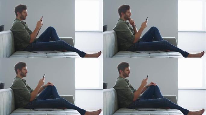英俊的大胡子男人坐在客厅的沙发上，在社交媒体上聊天滚动调情，使用蜂窝电话键入消息。自由职业者与朋友聊