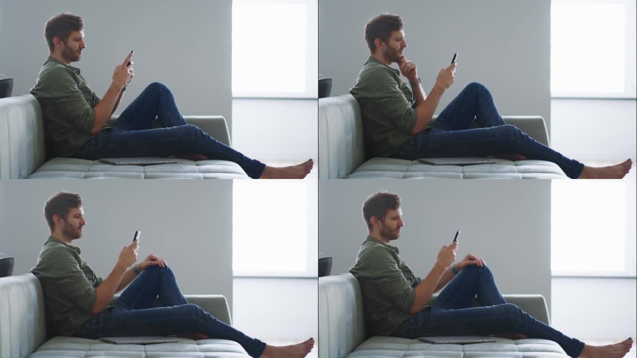 英俊的大胡子男人坐在客厅的沙发上，在社交媒体上聊天滚动调情，使用蜂窝电话键入消息。自由职业者与朋友聊