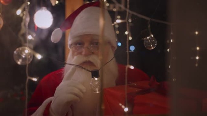 圣诞老人透过窗户和圣诞灯看着相机。圣诞老人带来了礼物