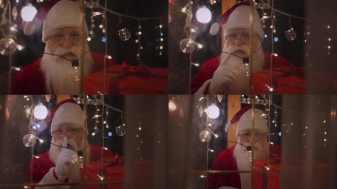 圣诞老人透过窗户和圣诞灯看着相机。圣诞老人带来了礼物
