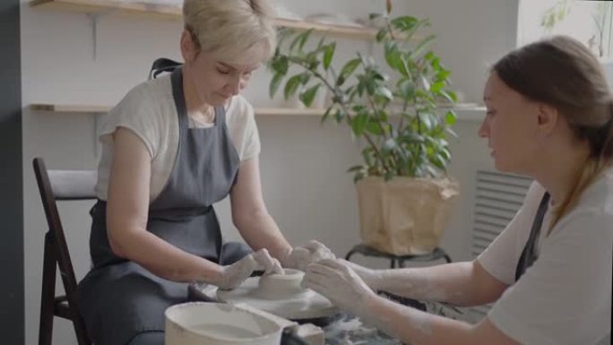 女大师将知识转移给一位在陶工轮上工作的老年妇女，并在她的工作室中以慢动作制作一杯陶瓷