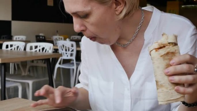 一名在咖啡馆穿着白衬衫的白人妇女在一张桌子上吃午餐，一只手拿着沙威玛，另一只手使用智能手机继续在线工
