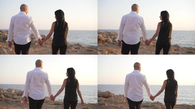 后视图充满爱心的快乐高加索夫妇手牵着手走向海岸悬崖。微笑的美女和英俊的男人享受约会漫步在慢动作分享。