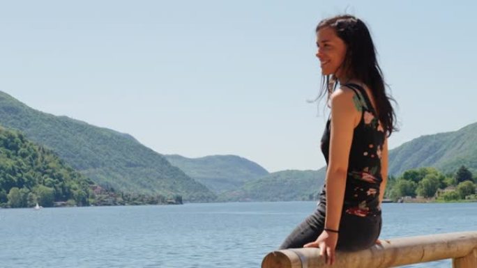 女人坐在木栏杆上，可以看到湖泊和绿色山脉的景色