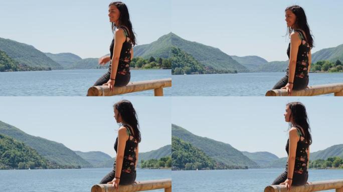 女人坐在木栏杆上，可以看到湖泊和绿色山脉的景色