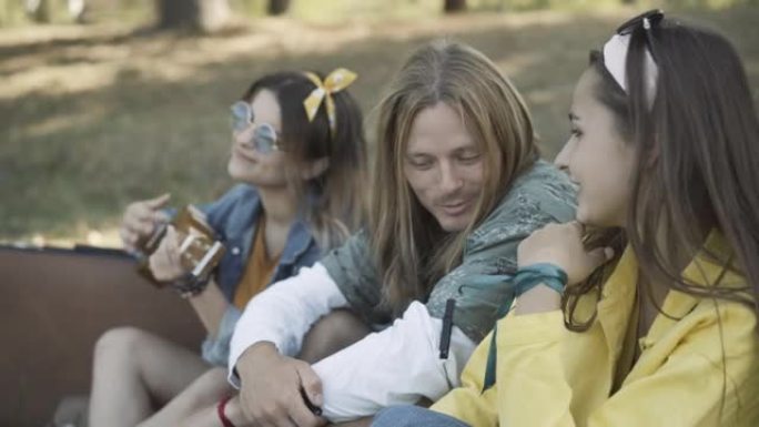 快乐放松的嬉皮士朋友坐在户外，聊天和玩夏威夷四弦琴的侧视图。积极的年轻高加索20世纪60年代男女在夏