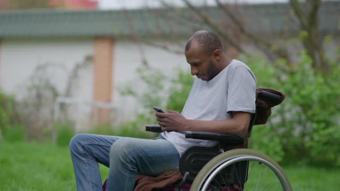 坐在轮椅上的瘫痪的非洲裔美国男子在慢动作的智能手机上浏览社交媒体的中景侧视图。集中残疾人在夏季户外花
