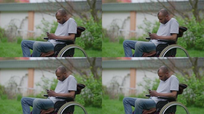 坐在轮椅上的瘫痪的非洲裔美国男子在慢动作的智能手机上浏览社交媒体的中景侧视图。集中残疾人在夏季户外花