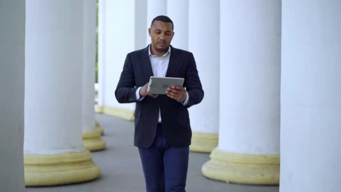 集中英俊的非洲裔美国商人穿着西装在平板电脑上上网，走在柱子之间。成功的年轻人在户外在线传递信息的肖像
