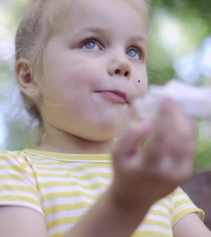 垂直视频: 小女孩用餐巾纸擦脸吃完后，桌上有奶昔。小女孩坐在公园长椅上，用纸巾擦脸的特写镜头。