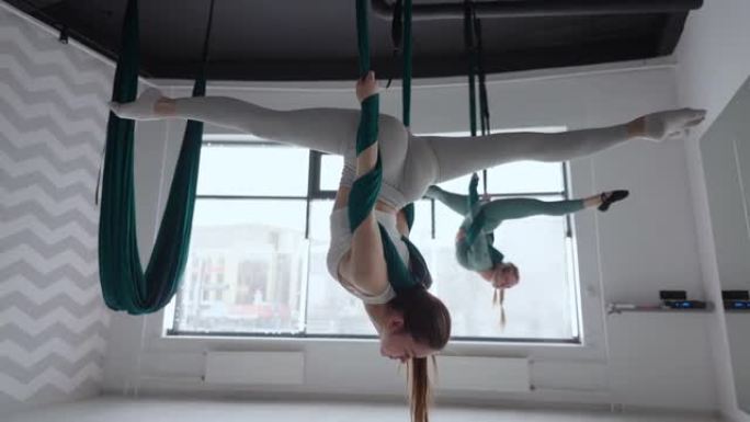 教练和年轻女子在健身房练习悬浮航空体操。在健身俱乐部的绿色吊床上进行空中瑜伽练习的两名年轻美丽的瑜伽