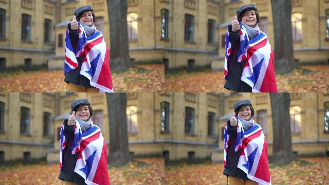 可爱的男生，露齿微笑，竖起大拇指，看着站在英国国旗的大学院子里的相机。自信聪明快乐的学生在校园户外摆