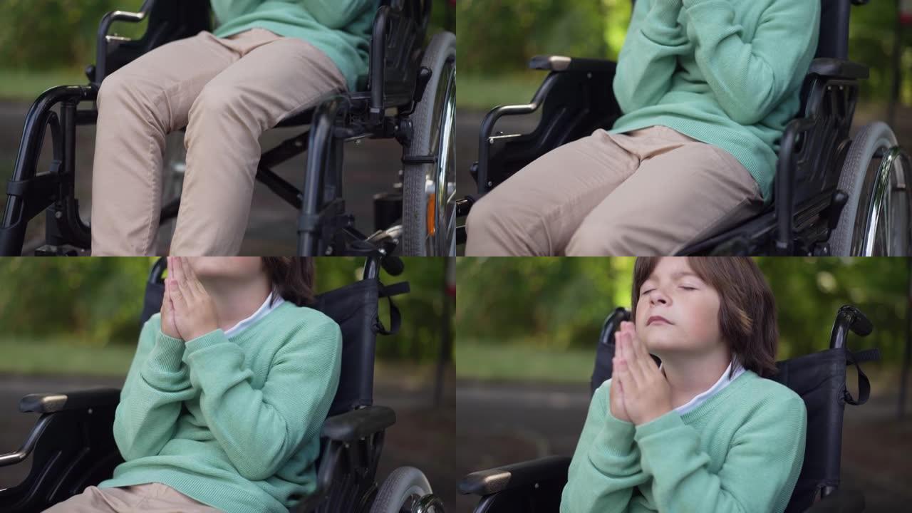 基座拍摄轮椅上的残疾儿童，闭着眼睛，双手紧握在户外祈祷。充满希望的高加索迷人男孩坐在夏季公园里向上帝