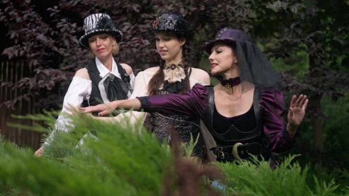 三个穿着蒸汽朋克服装的白人女人站在森林里触摸植物，用慢镜头说话。苗条美丽自信的女士肖像穿着万圣节服装