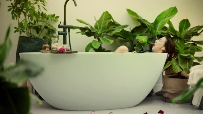 年轻女子在浴室内郁郁葱葱的绿色花园里洗泡泡浴。向手持淋浴唱歌