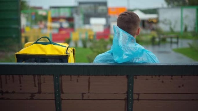 后视图年轻的送货员，黄色背包坐在城市街道的长凳上。穿着雨衣的白人男性快递员在雨后在镇上户外等待客户。