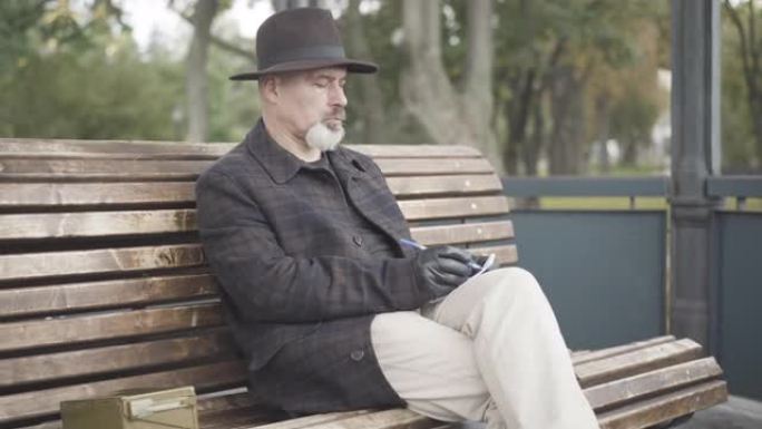 严肃的成年白人男子戴着优雅的帽子和皮手套坐在公园的长椅上写笔记。肖像专业私家侦探或卧底探员策划调查。