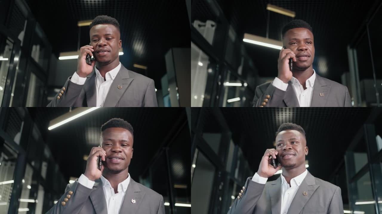 非洲商人在办公室内部说话智能手机。微笑的黑人商人在走廊里打电话给手机。友好的非洲裔美国人走在商务中心