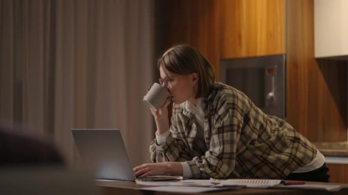 一位年轻漂亮的女商人在家工作，喝杯咖啡，看着站在桌子旁的笔记本电脑屏幕。查看邮件和喝咖啡。在家远程工