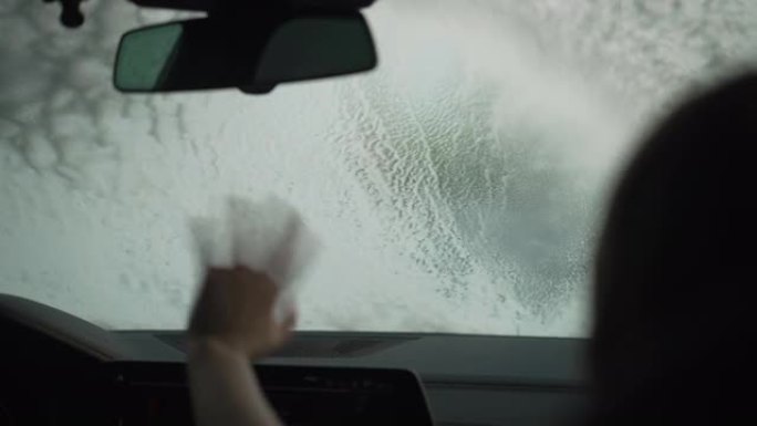 当男孩在车内挥舞时，挡风玻璃上覆盖着泡沫，用高压水冲走。在汽车洗车服务中玩耍的孩子的剪影。车辆维护概