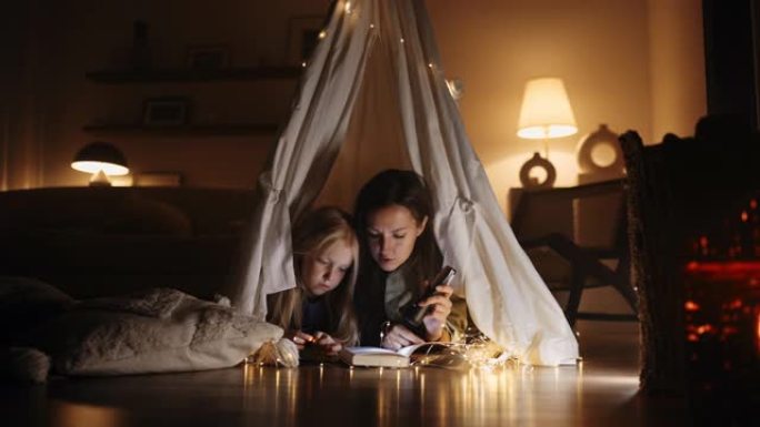 一个年轻的西班牙裔母女俩拿着手电筒在家里的帐篷里看书，上面装饰着房子的黑暗，神奇的装饰。母女舒适的氛
