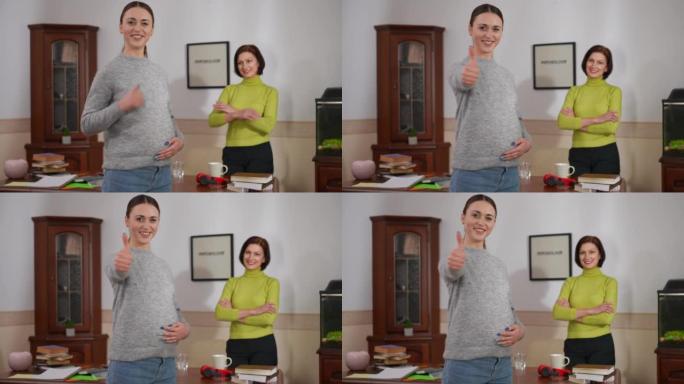 快乐的孕妇指着拇指抬头看着相机微笑，模糊的心理学家交叉双手站在背景下。积极满意的高加索患者和医生在室