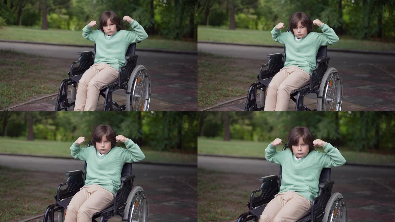 放大坐在轮椅上的白人残疾男孩，看着相机做着力量手势。自信的孩子在夏季公园户外摆姿势的肖像。残疾和动机