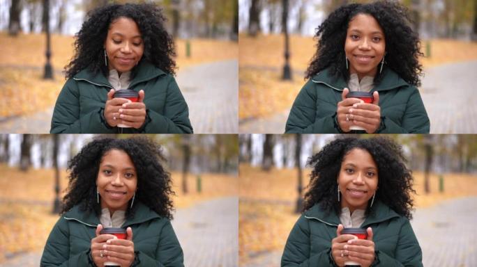 微笑迷人的年轻女子站在公园小巷，拿着咖啡杯看着相机的肖像。非裔美国人自信的女士在秋日户外摆姿势享受休
