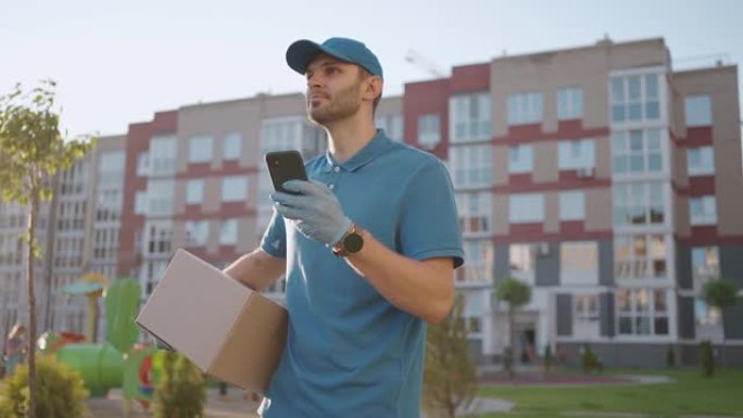 戴眼镜的邮递员携带包裹，并通过手机查看收货地址。搜索送货客户的地址。带帽和盒子的送货员