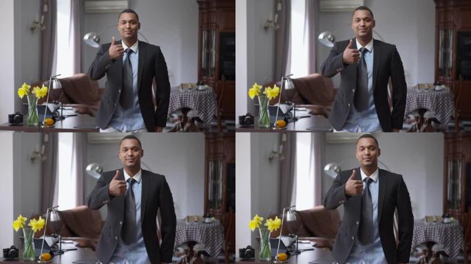 自信的年轻英俊的非洲裔美国男子站在家庭办公室的肖像，露出拇指微笑地看着相机。满意的成功经理在大流行封