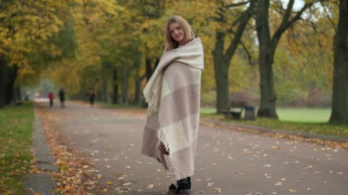 宽幅镜头自信的漂亮少女裹着毯子，站在小巷里看着相机。在金色的秋天户外微笑着摆姿势的高加索可爱的少年肖