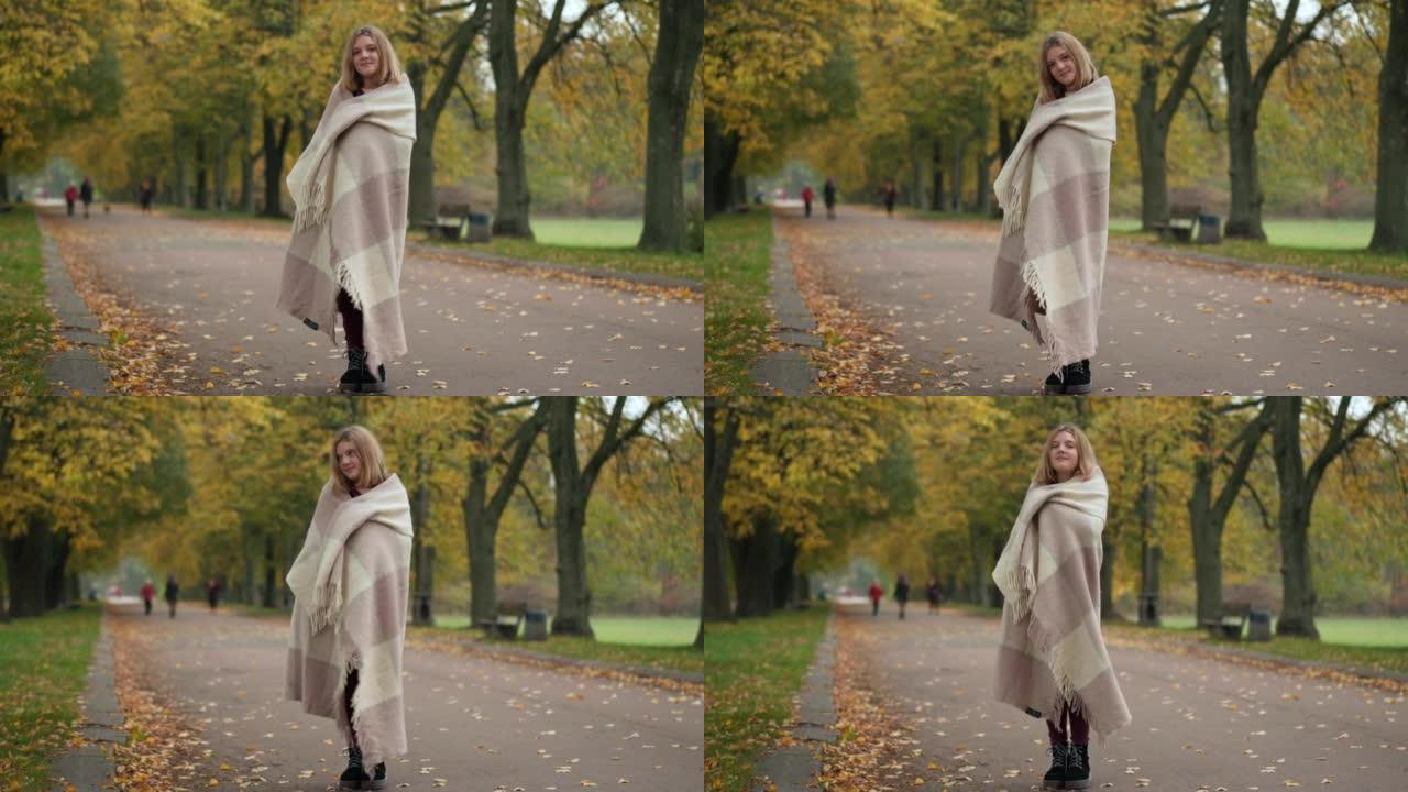 宽幅镜头自信的漂亮少女裹着毯子，站在小巷里看着相机。在金色的秋天户外微笑着摆姿势的高加索可爱的少年肖