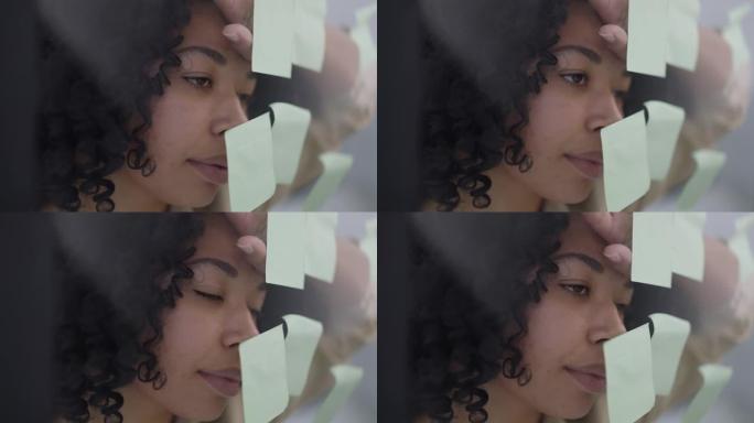 精疲力竭的非洲裔美国妇女的特写脸，用便签将额头放在玻璃门上。室内家庭办公室里超负荷工作的女商人的头像