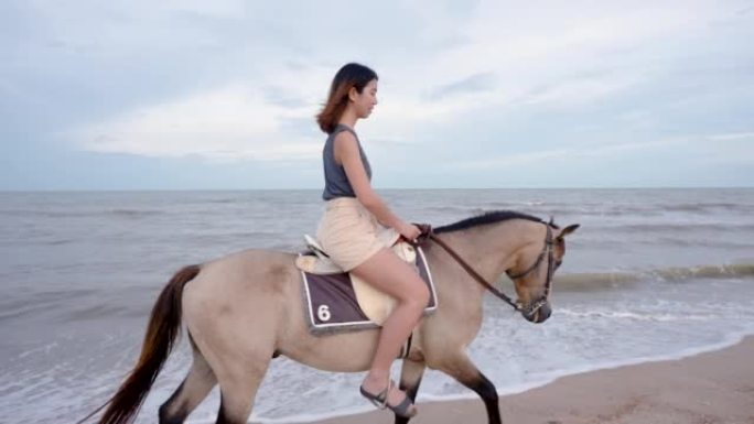 4K，一位年轻的亚洲妇女在太阳下山之前在沙滩上骑着马快乐地行走。游客来海滩放松，骑马，散步。